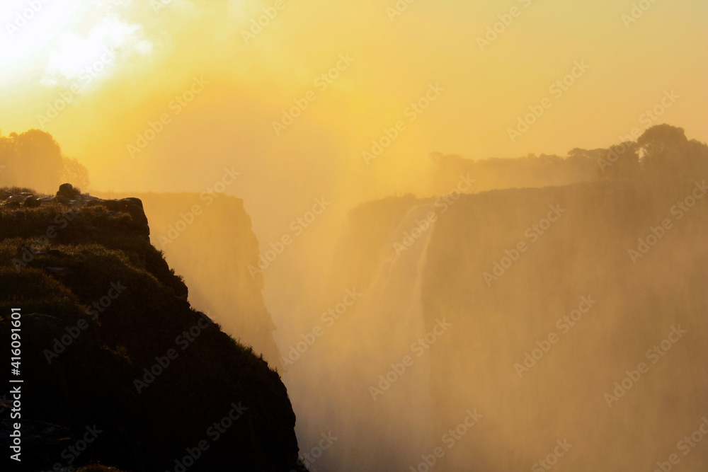 Sunrise over the mist and water dust of Victoria Falls in shades of red, orange and yellow.