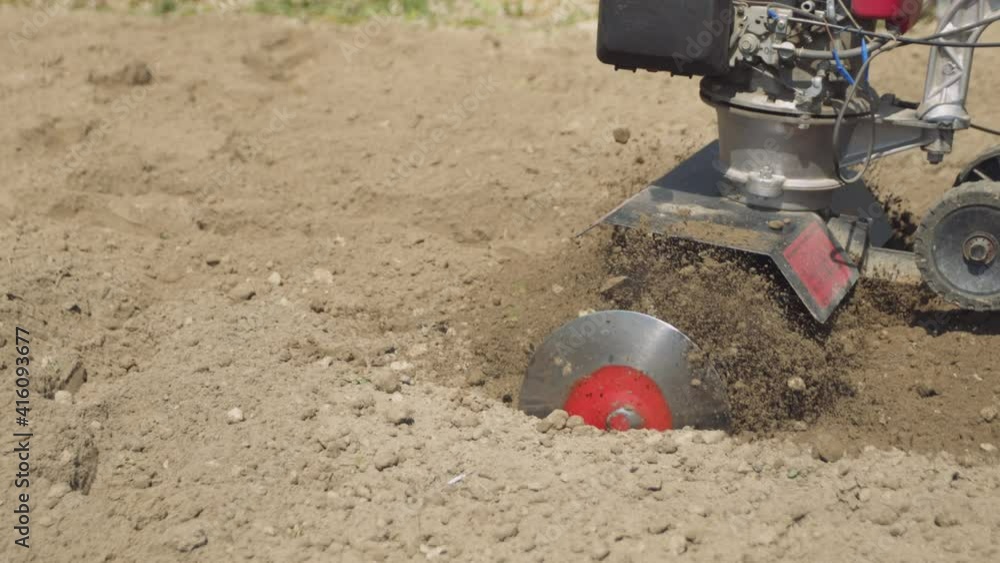 Farmer cultivates the soil in the garden using a tiller - motor cultivator. Man in hat and rubber boots. Footage 4k. Side view Farmer cultivates the soil in the garden using a tiller - motor cultivator. Man in hat and rubber boots. Footage 4k. Side view
