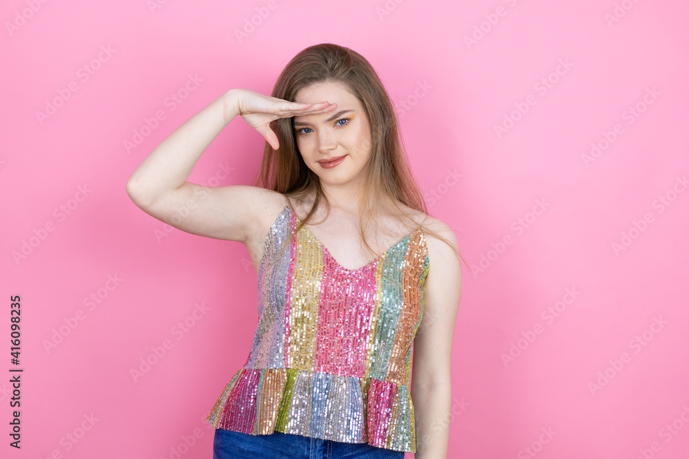Pretty redhead woman wearing a shiny top standing over pink background very happy and smiling looking far away with hand over head. searching concept. Pretty redhead woman wearing a shiny top standing over pink background very happy and smiling looking far away with hand over head. searching concept.