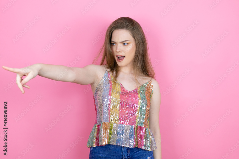 Pretty redhead woman wearing a shiny top standing over pink background pointing with finger surprised ahead, open mouth amazed expression, something on the front Pretty redhead woman wearing a shiny top standing over pink background pointing with finger surprised ahead, open mouth amazed expression, something on the front