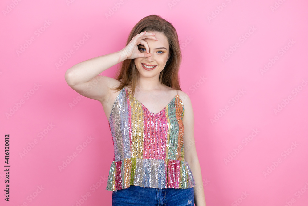 Pretty redhead woman wearing a shiny top standing over pink background doing ok gesture shocked with smiling face, eye looking through fingers Pretty redhead woman wearing a shiny top standing over pink background doing ok gesture shocked with smiling face, eye looking through fingers
