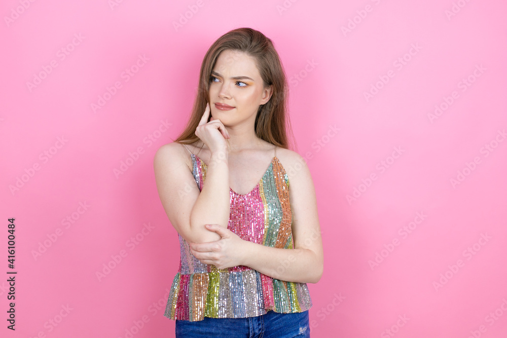 Pretty redhead woman wearing a shiny top standing over pink background with hand on chin thinking about question, pensive expression. smiling and thoughtful face. doubt concept. Pretty redhead woman wearing a shiny top standing over pink background with hand on chin thinking about question, pensive expression. smiling and thoughtful face. doubt concept.