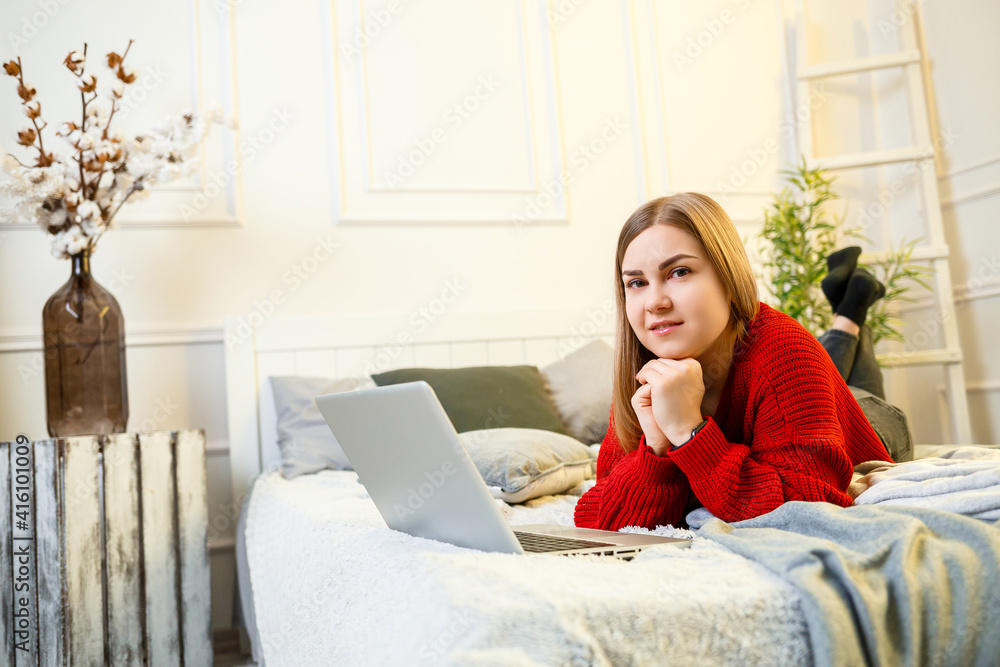 Young beautiful woman in a red sweater typing on a laptop sitting on a bed in a bright room. Working remotely in a home atmosphere during quarantine