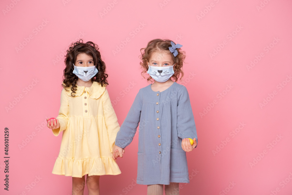 two girls in protective mask with colored eggs holding hands on pink background. Covid easter concept