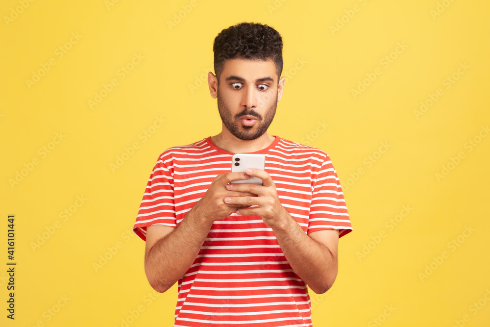 Shocked bearded man in striped t-shirt texting on smartphone looking at display with big eyes, surprised with information or news. Indoor studio shot isolated on yellow background Shocked bearded man in striped t-shirt texting on smartphone looking at display with big eyes, surprised with information or news. Indoor studio shot isolated on yellow background