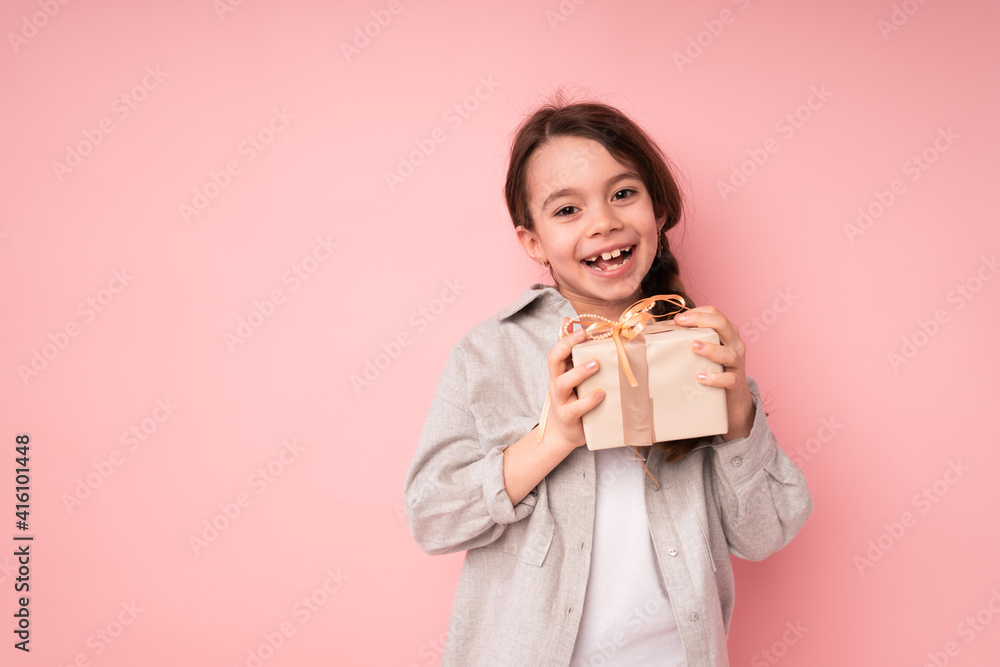 Little happy girl stands on a bright background and smiles holding a wrapped gift in her hands Little happy girl stands on a bright background and smiles holding a wrapped gift in her hands