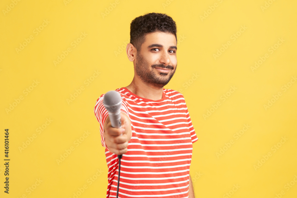 Bearded male reporter in striped t-shirt holding microphone, making interview and asking opinion, discussing important topics. Indoor studio shot isolated on yellow background Bearded male reporter in striped t-shirt holding microphone, making interview and asking opinion, discussing important topics. Indoor studio shot isolated on yellow background