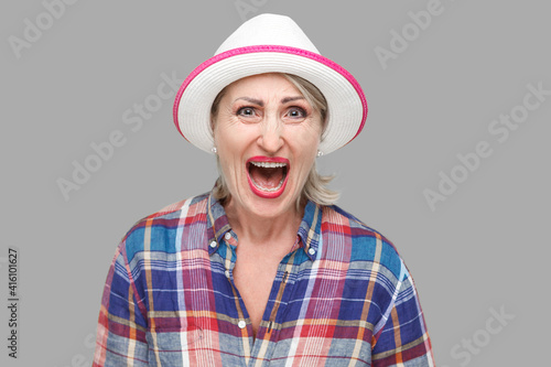 Portrait of angry or shocked modern stylish mature woman in casual style with white hat standing and looking at camera and screaming. indoor studio shot isolated on gray background.