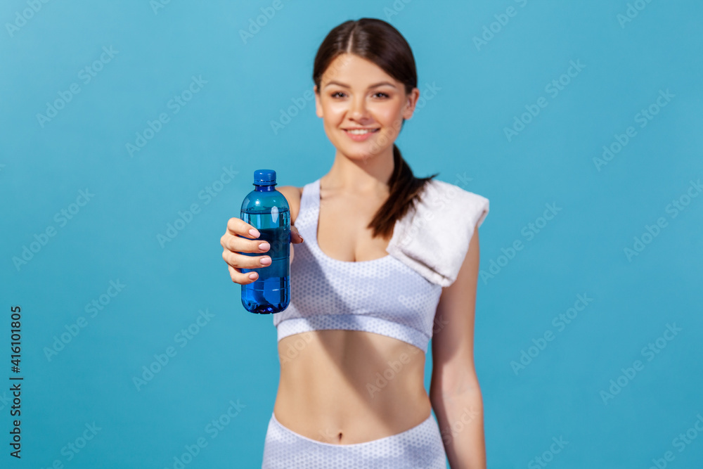 Healthy smiling woman athlete in white sportswear with towel on shoulder holding and showing blue bottle with mineral water, restoring electrolytes. Indoor studio shot isolated on blue background Healthy smiling woman athlete in white sportswear with towel on shoulder holding and showing blue bottle with mineral water, restoring electrolytes. Indoor studio shot isolated on blue background