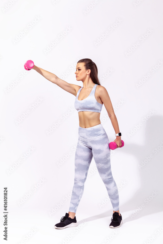Full length side view athletic woman in white sports top and tights raising hands with dumbbells, pumping up muscles and developing strength. Indoor studio shot isolated on gray background Full length side view athletic woman in white sports top and tights raising hands with dumbbells, pumping up muscles and developing strength. Indoor studio shot isolated on gray background