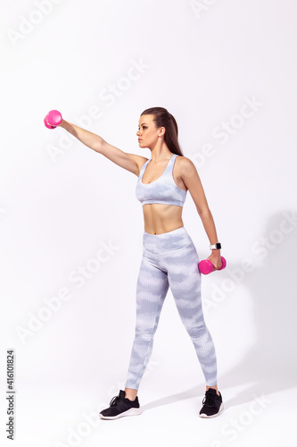 Full length side view athletic woman in white sports top and tights raising hands with dumbbells, pumping up muscles and developing strength. Indoor studio shot isolated on gray background