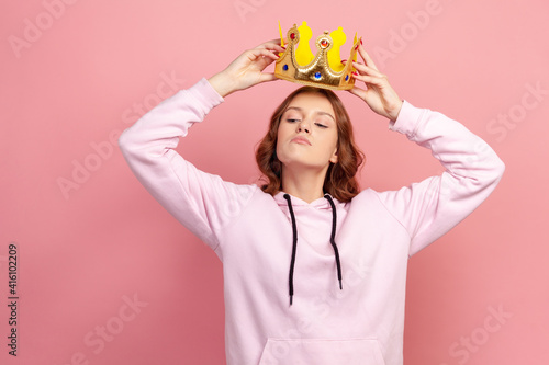 Tapeta Portrait of selfish curly haired teenage girl in hoodie holding gold crown over head, looking with arrogance, privileged status