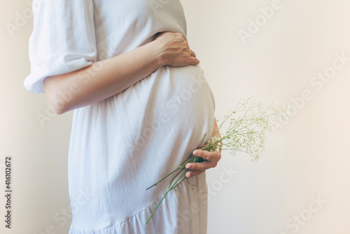 Pregnant woman in white dress holding belly and flowers with her hands.
