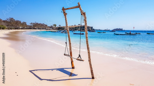 A swing placed on the seashore of Pink Beach, Lombok, Indonesia. The swing has very simple wood construction. Waves gently wash the pillars of it. In the back there are few boats anchored in the bay.