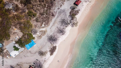 A drone shot of idyllic Pink Beach on Lombok, Indonesia. Sea is calm, shining with many shades of blue. Beauty in the nature. Unspoiled, hidden gem.There are few houses along the seashore. © Chris
