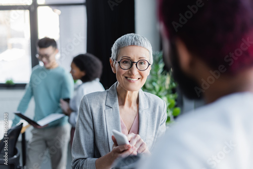 Cheerful businesswoman with smartphone talking to african american colleague