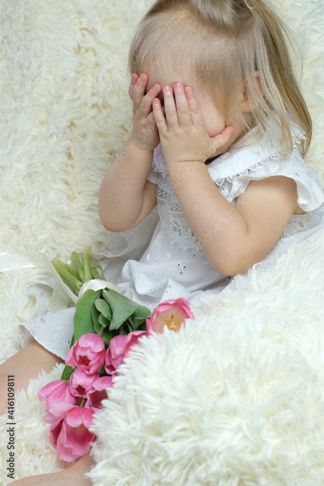 girl of two years old, blonde is sitting in the room on the couch with a bouquet of pink tulips, covered her eyes with her hands, concept small woman, birthday, happy childhood girl of two years old, blonde is sitting in the room on the couch with a bouquet of pink tulips, covered her eyes with her hands, concept small woman, birthday, happy childhood