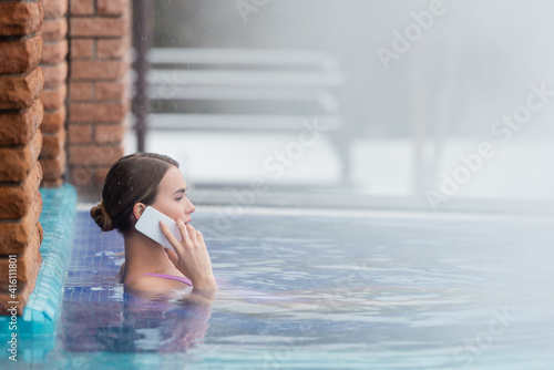 side view of woman in swimsuit talking on smartphone while bathing in outdoor swimming pool