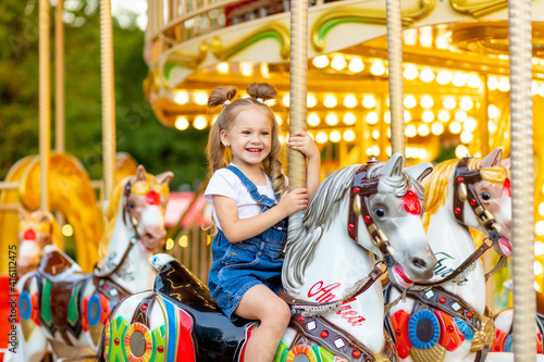 Fotografering happy baby girl rides a carousel on a horse in an amusement Park in summer
