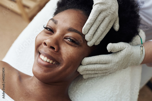 Facial massage with gloves with microcurrents. Microcurrent therapy. Cosmelotologist makes stimulating facial massage with electric gloves. Young happy black woman on cosmetic procedures. Skin care