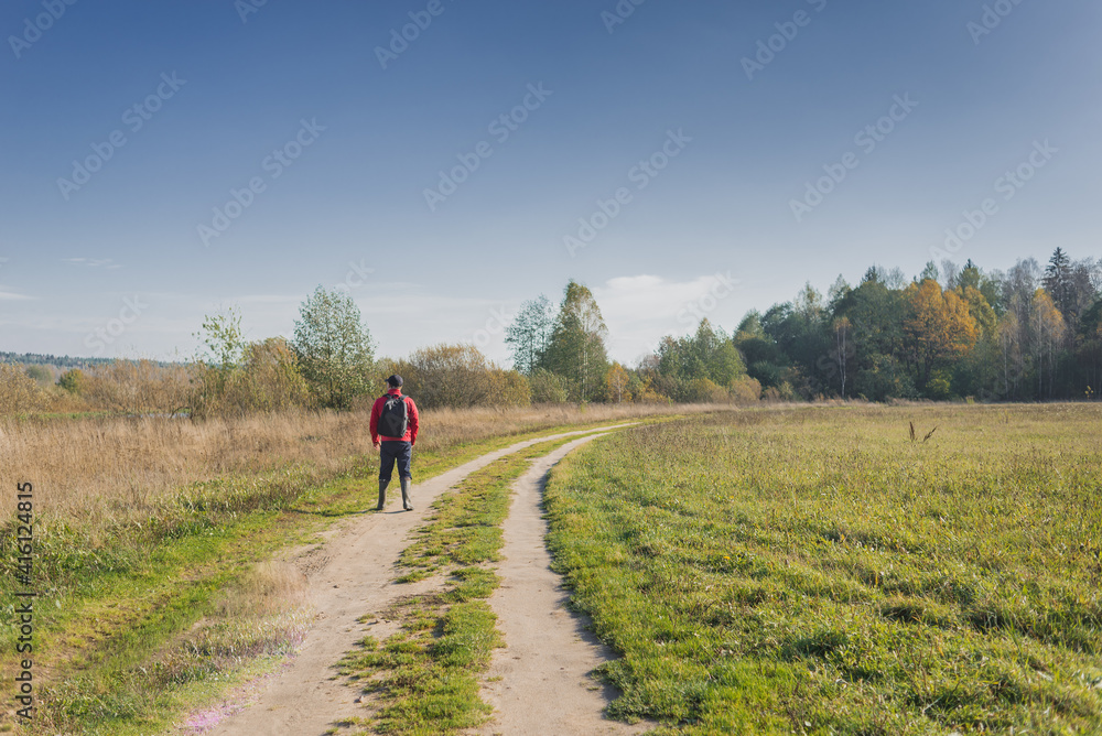 Traveler walks on the road in nature. Man in a red jacket with a backpack walks along a country road on a sunny day.
