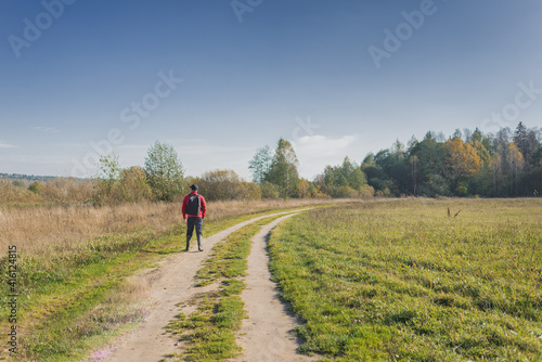 Traveler walks on the road in nature. Man in a red jacket with a backpack walks along a country road on a sunny day. © alexey351