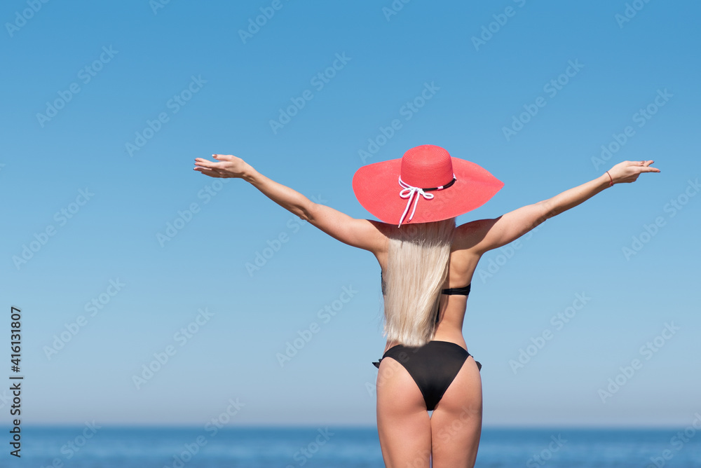 a slender blonde in a black swimsuit and a red hat stands on the beach and raises her hands to the sky, enjoying the sun. travel and vacation concept. sports woman on the background of the sea. a slender blonde in a black swimsuit and a red hat stands on the beach and raises her hands to the sky, enjoying the sun. travel and vacation concept. sports woman on the background of the sea.