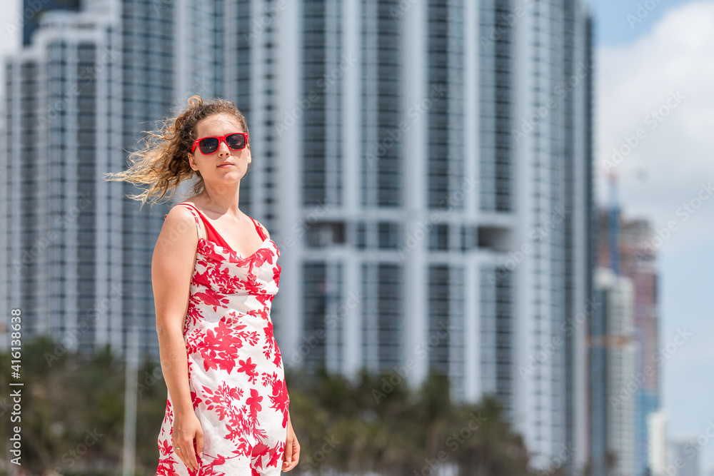 Fototapeta premium Portrait of young woman hipster millennial on beach on sunny day with red sunglasses in Miami, Florida with skyscrapers hotels apartments condo building in background