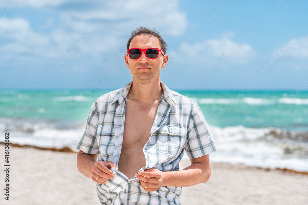 Young man with hands undressing unbuttoning shirt on beach on sunny day with red sunglasses in Miami, Florida with ocean waves in background