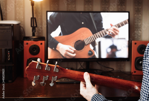 Man in a shirt practice play acoustic guitar in front of his computer monitor online staying home during quarantine and forced self-isolation due to coronavirus. Guitar neck close-up