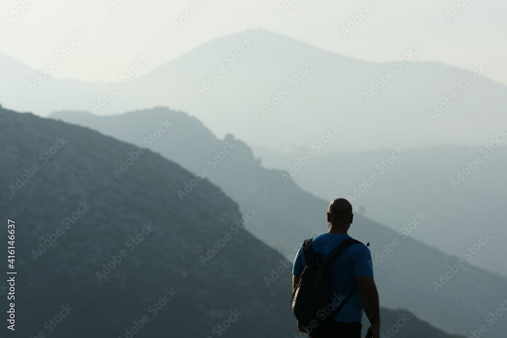 Hiker in a blue shirt and black pants, with his back turned, contemplating a beautiful landscape at sunset, with sunny mountains in the background. Hiker in a blue shirt and black pants, with his back turned, contemplating a beautiful landscape at sunset, with sunny mountains in the background.
