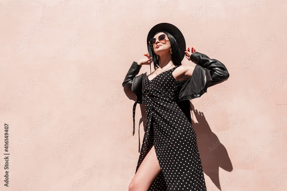 Fashion glamorous young woman in stylish sunglasses in leather jacket in beautiful dress straightens hat on sunny day. Lovely trendy girl in elegant black summer clothes enjoys sunlight outdoors. - obrazy, fototapety, plakaty 