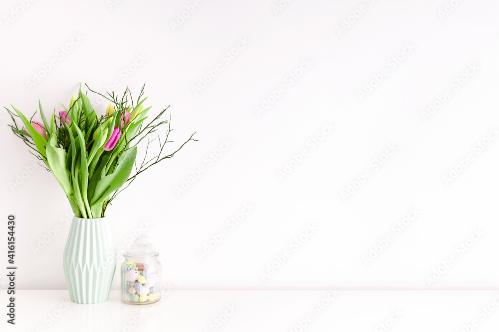 Bouquet of multicolored unopened tulips in light green ceramic vase and Easter candy jar filled with eggs on white wall background. Home interior decoration concept. Place for text.