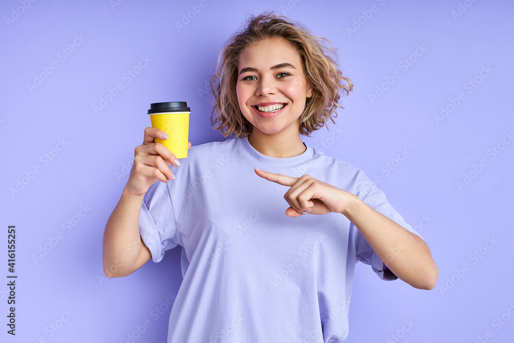 female with cup of coffee isolated on purple background, young short-haired woman in casual wear pointing index finger at yellow cup, smile. female with cup of coffee isolated on purple background, young short-haired woman in casual wear pointing index finger at yellow cup, smile.