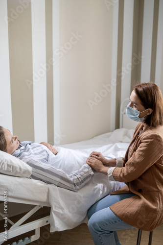 Young female in casualwear and protective mask holding hand of her sick friend