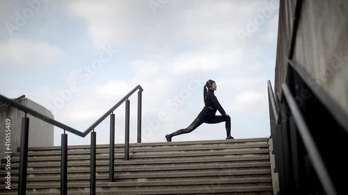 Woman in black sportswear doing stretching exercise outside in urban environment in the morning before a jogging session. Sports and urban concept.