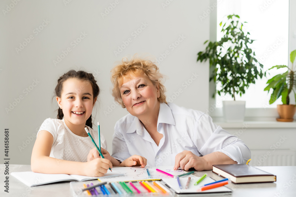 Happy grandchild with grandmother having fun, drawing colored pencils, sitting together at home, laughing preschool girl with smiling grandma painting picture, playing with granddaughter Happy grandchild with grandmother having fun, drawing colored pencils, sitting together at home, laughing preschool girl with smiling grandma painting picture, playing with granddaughter
