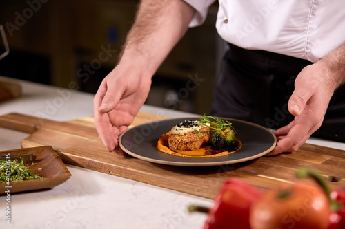 delicious dish on black plate represented by professional cropped cook in apron, plate on wooden board, fresh yummy meal