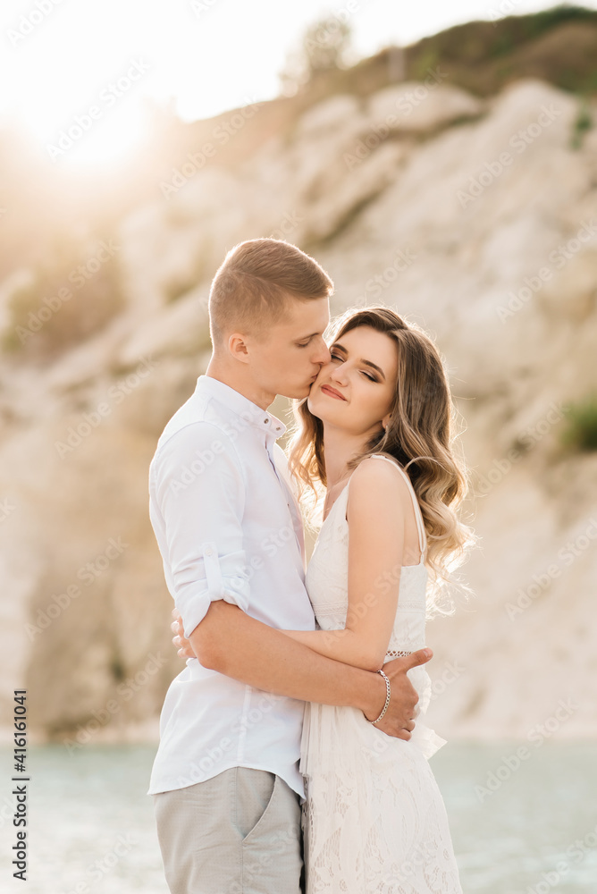 Fototapeta premium A beautiful young couple in love, a man and a woman embrace, kiss near a blue lake and sand at sunset. Vacation at the sea on the beach, sunset, morning, honeymoon, tropics, ocean, girl, relationship.