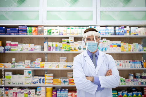 Portrait of pharmacist wearing face shield and white coat standing in pharmacy store during corona virus pandemic.