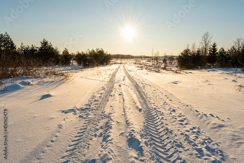 The trail from the tractor in the snow passes through the field to the horizon. Winter landscape in the evening with clear blue sky
