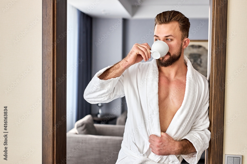 male in bathrobe begins the day with cup of hot tea or coffee, stand posing near the door, looking at side