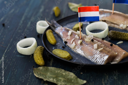 Traditional Dutch food herring fish with pickled cornichons and onion on dark blue ceramic plate and wooden background with flags. New season of herring called 