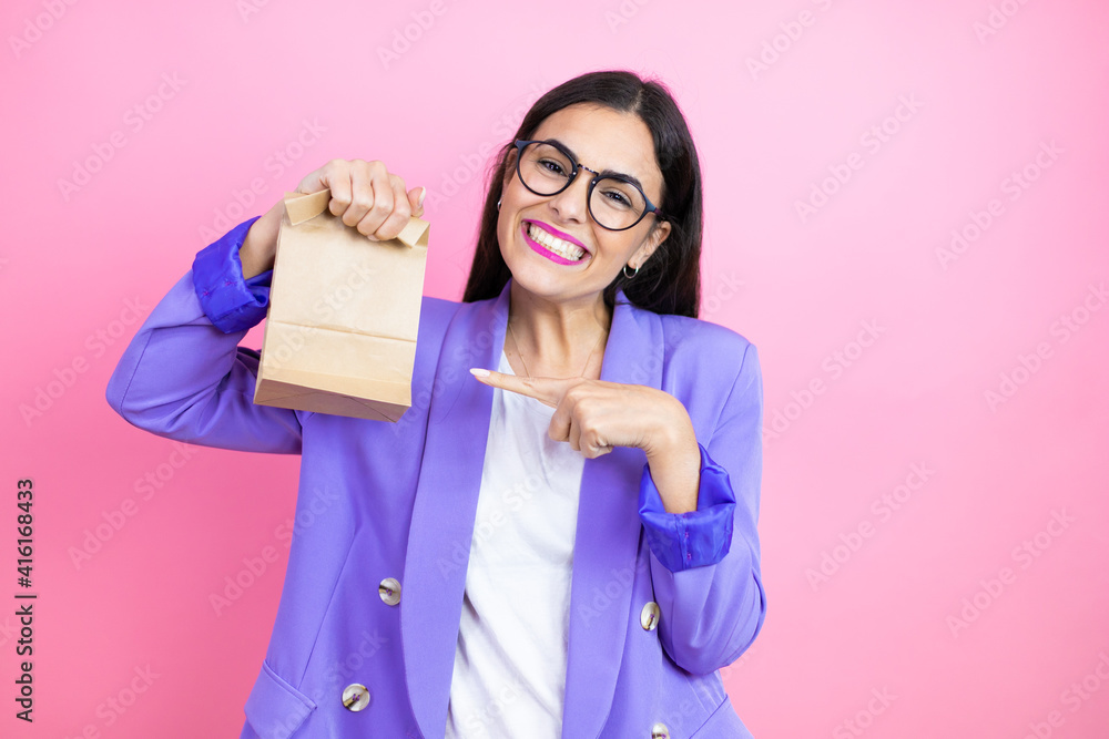 Young business woman wearing purple jacket over pink background smiling, surprised and pointing paper bag in her hand Young business woman wearing purple jacket over pink background smiling, surprised and pointing paper bag in her hand