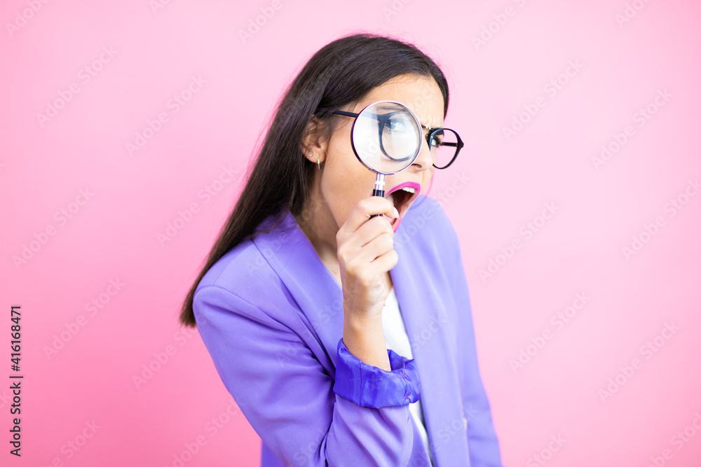 Young business woman wearing purple jacket over pink background surprised looking through a magnifying glass Young business woman wearing purple jacket over pink background surprised looking through a magnifying glass