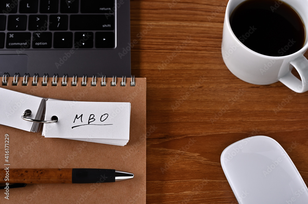 On the desk there is a laptop, a cup of coffee, and a word book with the word MBO written on it. It was an abbreviation for the financial term Management by Objectives. On the desk there is a laptop, a cup of coffee, and a word book with the word MBO written on it. It was an abbreviation for the financial term Management by Objectives.