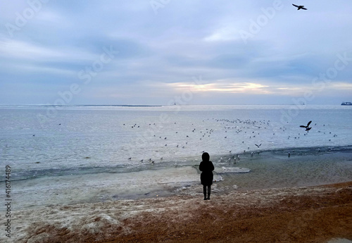 Silhouette of a woman in winter clothes on the background of the frozen sea. Winter evening on the beach, seagulls, pigeons. Cloudy sky. There is a breakwater on the horizon.