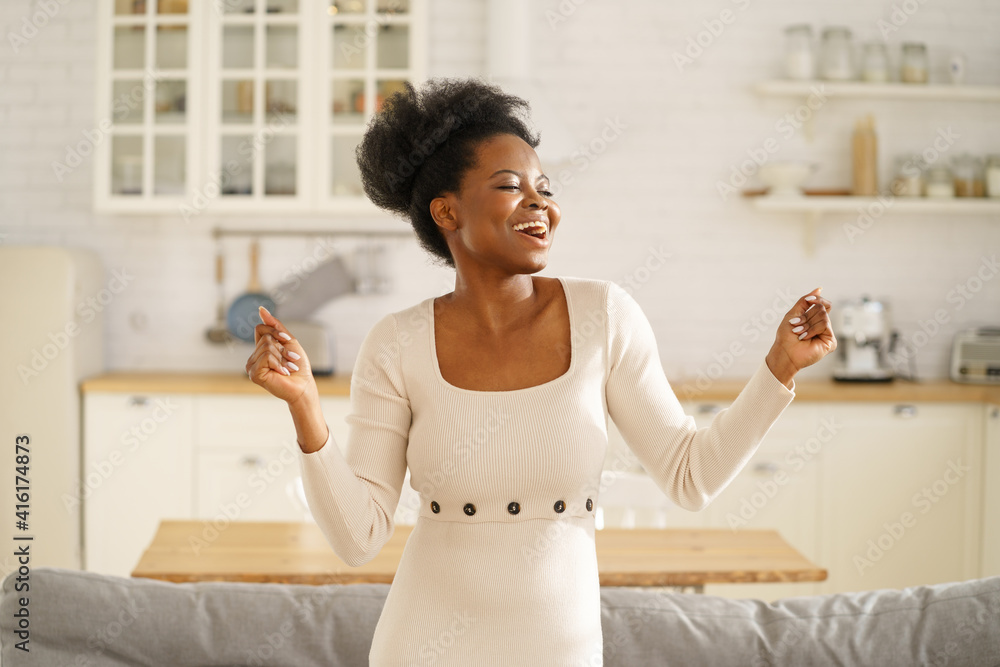Happy African American millennial woman wear fashion summer dress dancing, sings her favorite song, enjoying listening to music in living room. Carefree Black girl having fun alone on weekend at home. Happy African American millennial woman wear fashion summer dress dancing, sings her favorite song, enjoying listening to music in living room. Carefree Black girl having fun alone on weekend at home.