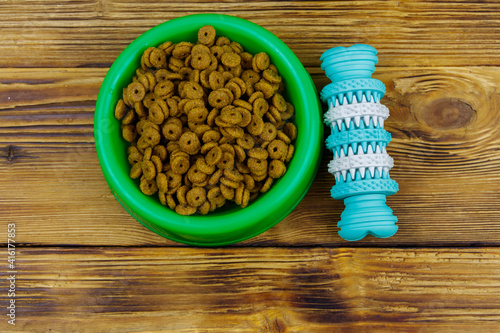 Dog toy and dry dog food in green plastic bowl on wooden background. Top view. Dog care concept