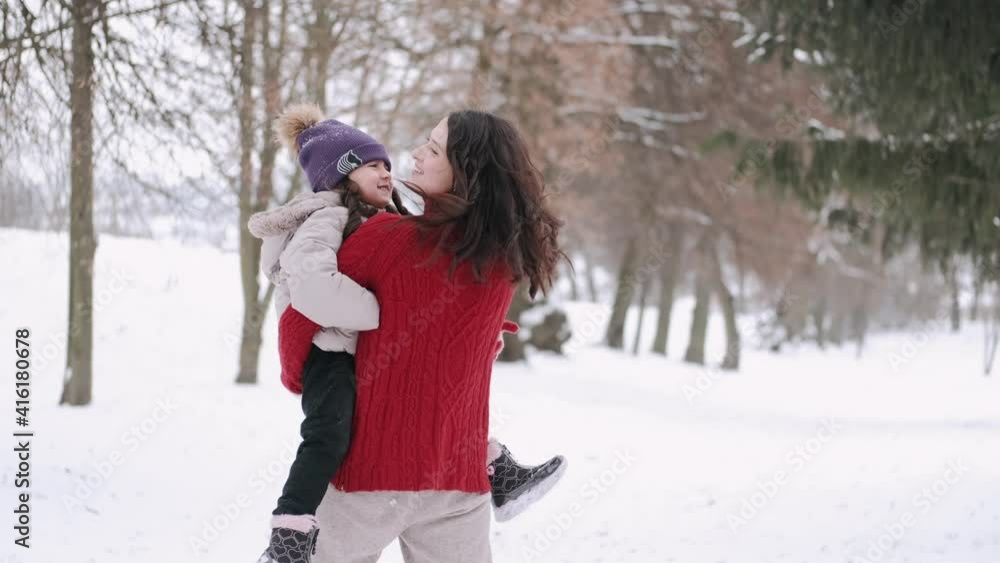 Little daughter in overalls and young adult mother walking on white first snow at park. Spending time together in beautiful winter day. Enjoying peaceful stroll. Little daughter in overalls and young adult mother walking on white first snow at park. Spending time together in beautiful winter day. Enjoying peaceful stroll.